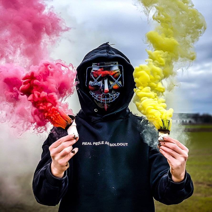 Hooded figure in neon gas mask holding pink and yellow smoke bombs outdoors HD phone wallpaper
