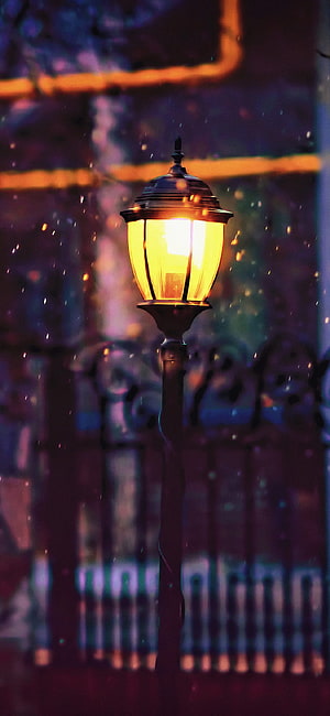 Glowing vintage street lamp beside ornate iron fence at night with falling snow HD phone wallpaper