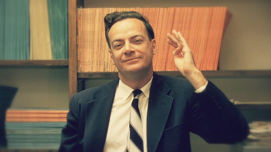 Richard Feynman in a dark suit and striped tie waving in front of a bookshelf HD wallpaper