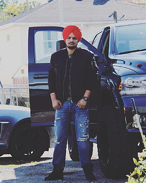 Sidhu Moose Wala in red turban and black jacket standing beside a lifted truck HD phone wallpaper