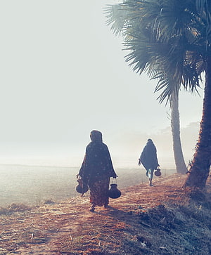 Two women carrying water pots walk along a rural path in Bangladesh during winter morning HD phone wallpaper