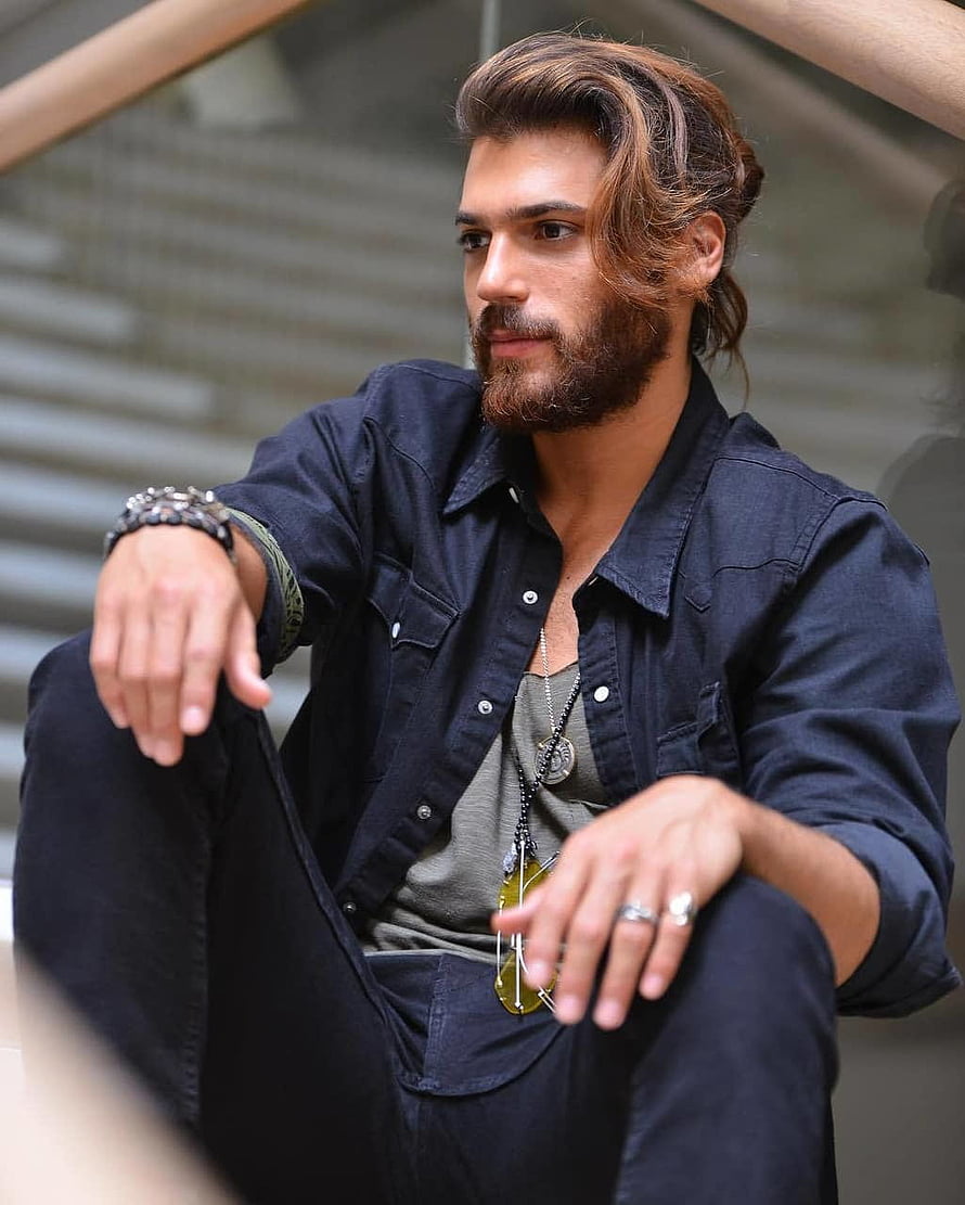 Can Yaman in a dark denim shirt with layered necklaces and a full beard, seated in a modern architectural setting HD phone wallpaper