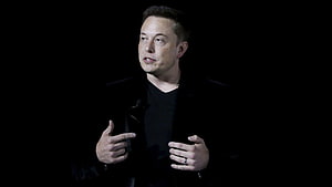 Elon Musk Speaking on Stage in Black Attire Against Dark Background HD wallpaper