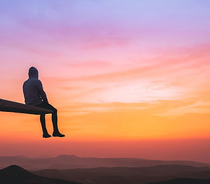 Silhouette of a hooded man sitting on a wooden ledge overlooking a mountain range at sunset HD wallpaper