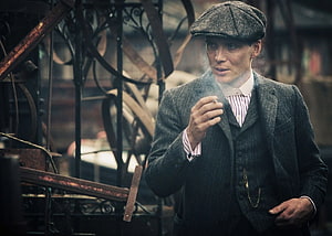 Tommy Shelby in vintage suit and flat cap smoking a cigarette in a dim industrial setting HD wallpaper