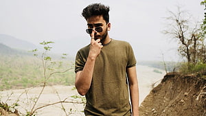 Elvish Yadav in olive t-shirt and sunglasses standing outdoors near a river with hazy hills in the background HD wallpaper