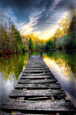 Weathered wooden dock extending into a calm lake at golden hour with lush green trees and dramatic sky reflections HD phone wallpaper