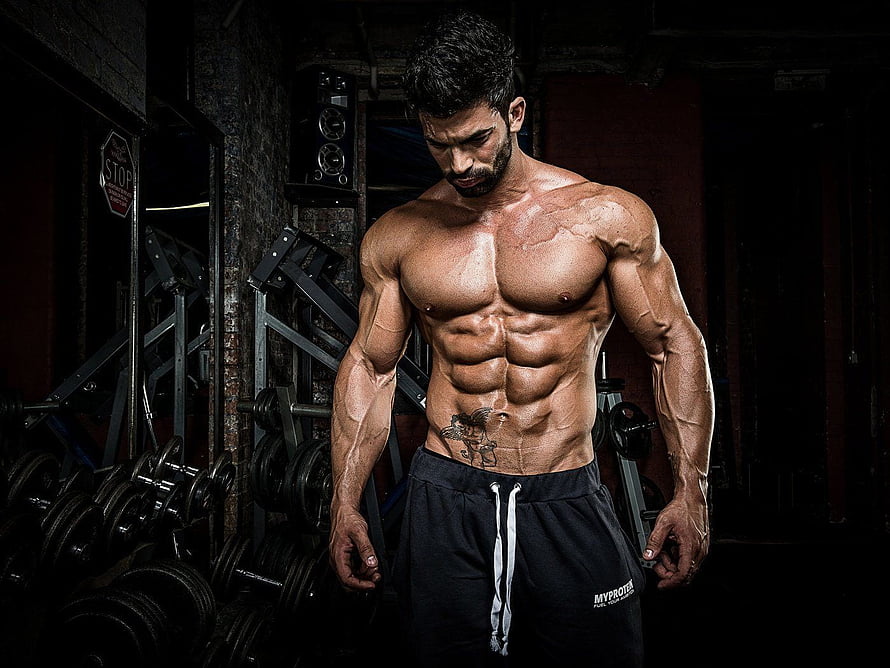 Muscular shirtless man with tattoo posing in a dark gym with weight plates HD wallpaper
