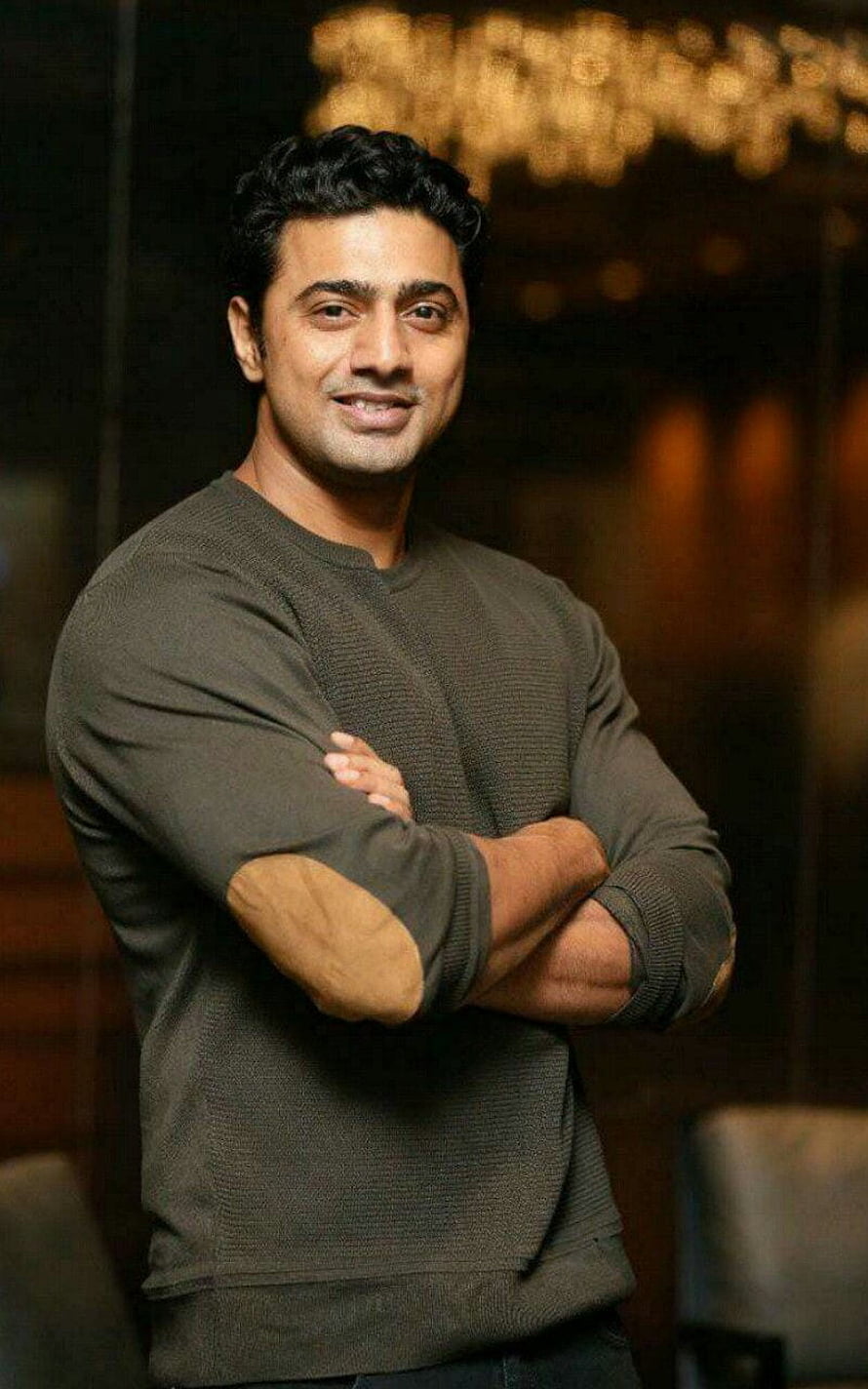 Dev Adhikari in a dark olive sweater with arms crossed against a warm bokeh background HD phone wallpaper