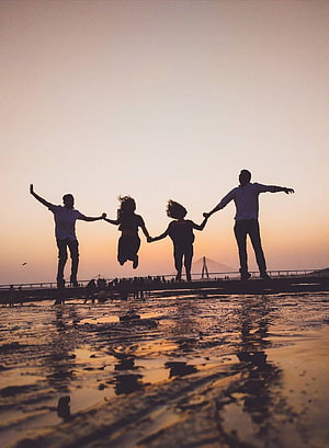 Silhouetted friends jumping together on a wet beach at sunset with a bridge in the distance HD phone wallpaper