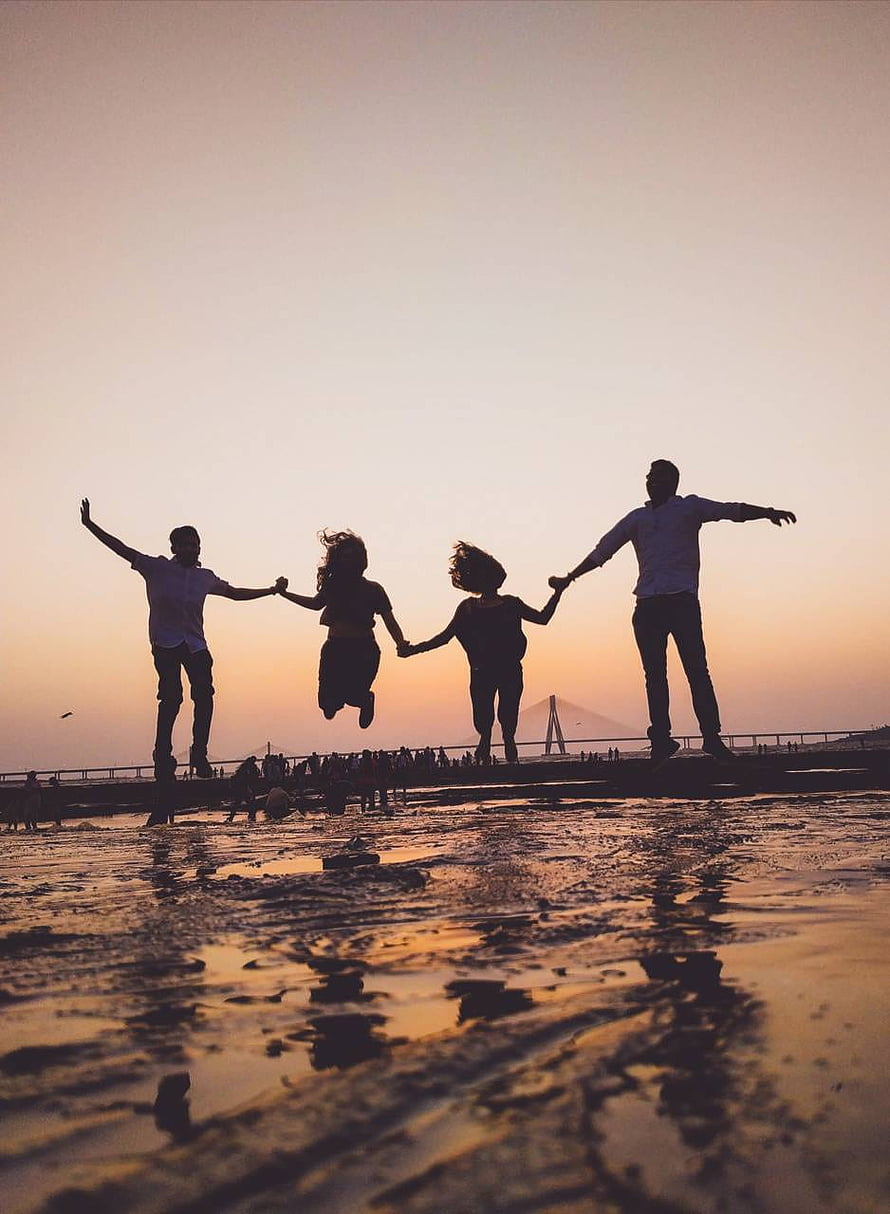 Silhouetted friends jumping together on a wet beach at sunset with a bridge in the distance HD phone wallpaper