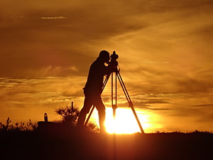 Silhouette of a surveyor operating a theodolite on a tripod against a golden sunset sky HD wallpaper