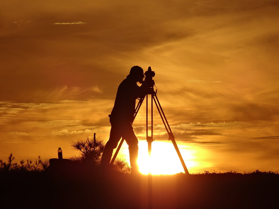 Silhouette of a surveyor operating a theodolite on a tripod against a golden sunset sky HD wallpaper