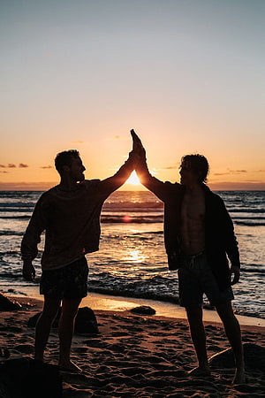 Two friends high-fiving on a sandy beach at sunset with ocean waves in the background HD phone wallpaper