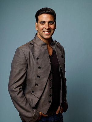 Akshay Kumar smiling in a grey double-breasted jacket against a blue studio backdrop HD phone wallpaper