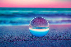 Glass sphere on a sandy beach reflecting a vibrant pink and teal ocean sunset HD wallpaper