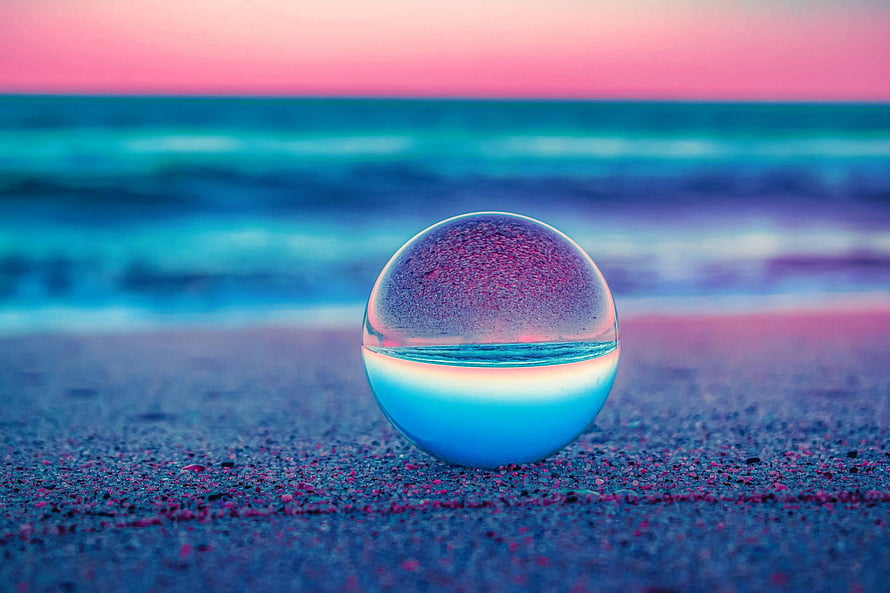 Glass sphere on a sandy beach reflecting a vibrant pink and teal ocean sunset HD wallpaper