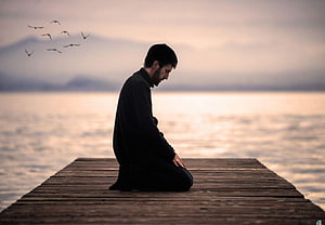 Man in Traditional Attire Kneeling in Prayer on a Wooden Dock at Dusk HD wallpaper