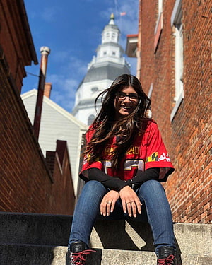Young woman in red Maryland jersey sitting on stone steps with historic church steeple in background, Mia Khalifa HD phone wallpaper