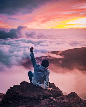 Man Sitting on Mountain Peak with Raised Fist Above Sea of Clouds at Sunset HD phone wallpaper