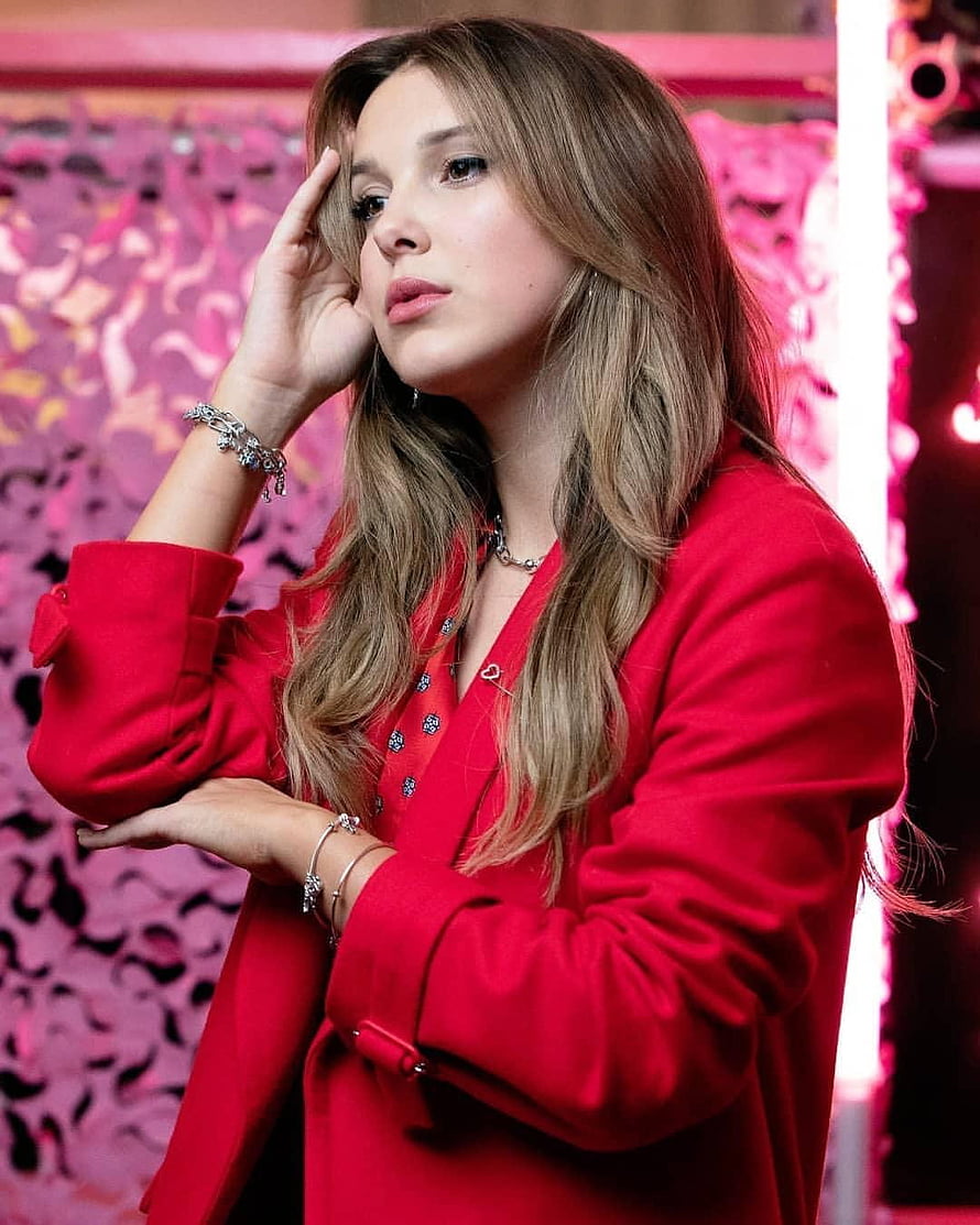 Millie Bobby Brown in a striking red coat with silver jewelry against a pink patterned backdrop HD phone wallpaper