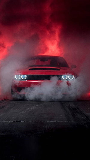 Red Dodge Challenger SRT Hellcat emerging from thick smoke with glowing headlights in a dark studio setting HD phone wallpaper
