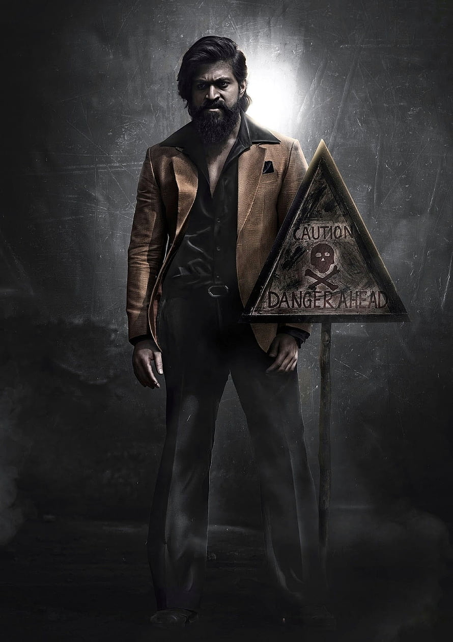 Rocky from KGF Chapter 2 in a tan suit beside a danger sign in a dark gritty setting HD phone wallpaper