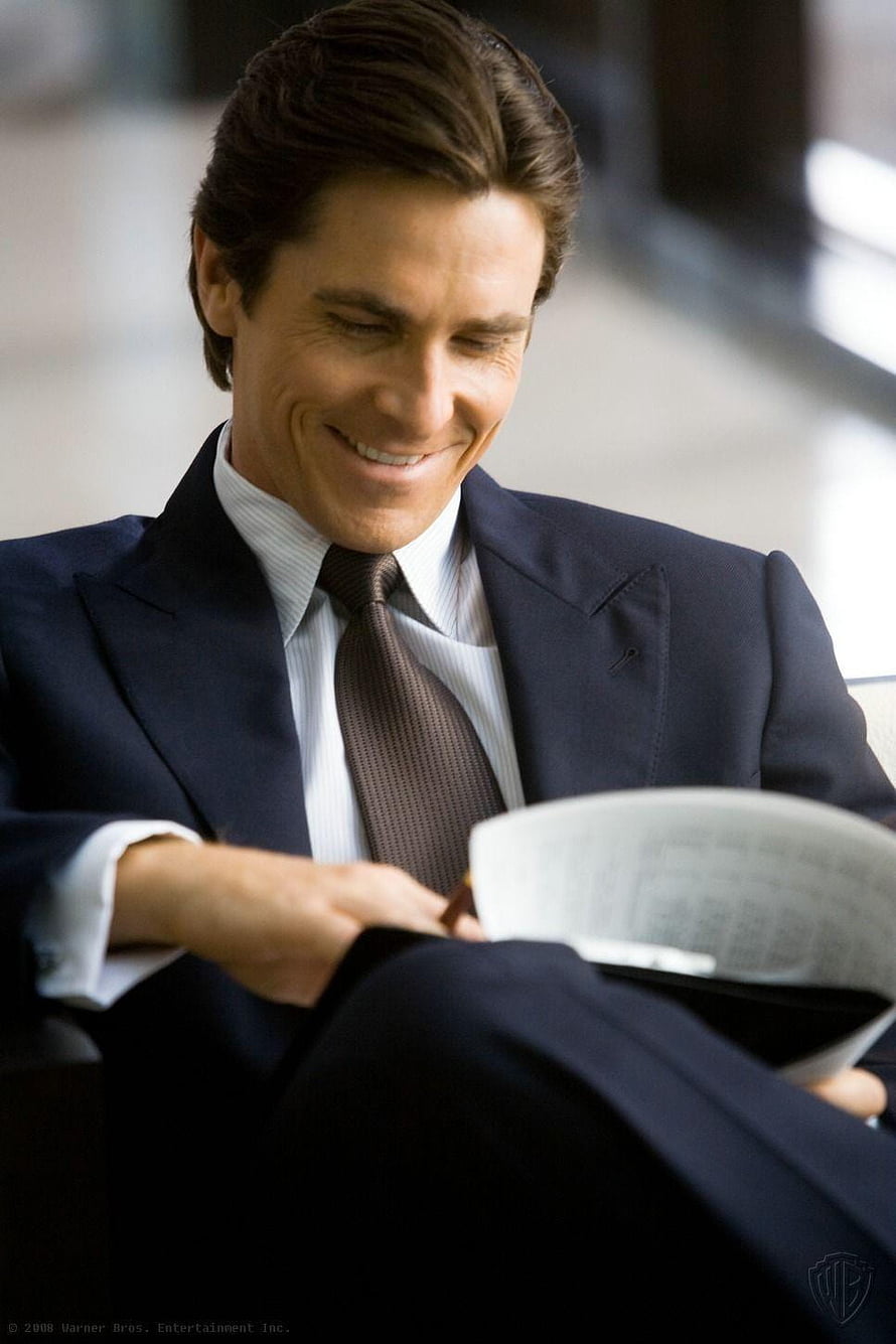Christian Bale as Bruce Wayne in a navy suit reading a newspaper HD phone wallpaper