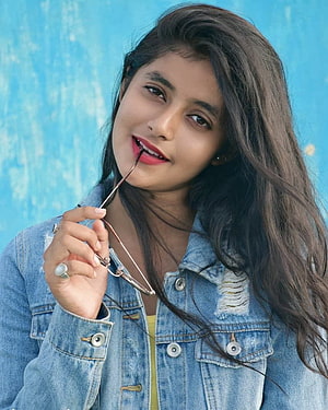Sanchita Basu in a distressed denim jacket holding glasses against a blue wall HD phone wallpaper