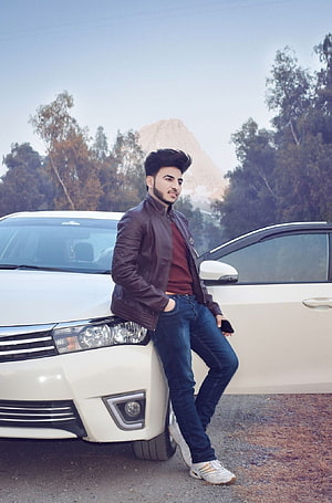 Young man in brown leather jacket leaning against white sedan outdoors HD phone wallpaper