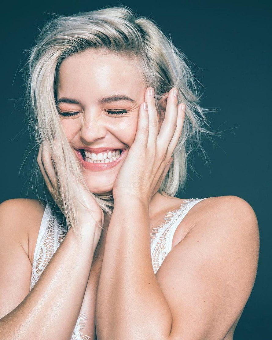 Anne Marie smiling portrait with hands on face in white lace top against dark teal background HD phone wallpaper