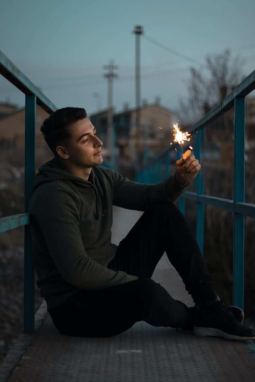 Young man sitting on a bridge at dusk holding a lit sparkler in a contemplative pose HD phone wallpaper