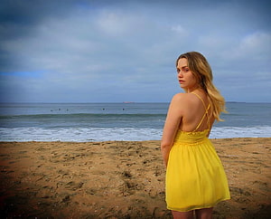 Mia Malkova in a yellow dress standing on a sandy beach with ocean waves and cloudy sky HD wallpaper