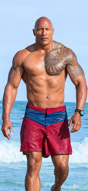 Dwayne Johnson shirtless on the beach in red swim trunks with ocean waves behind him HD phone wallpaper