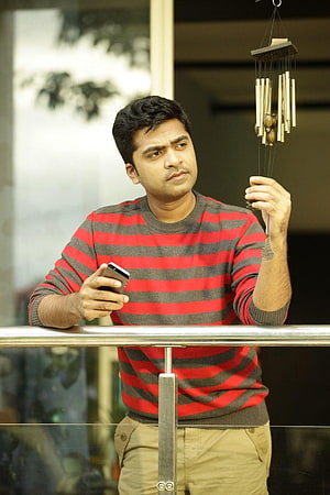 Simbu in red and grey striped sweater holding a wind chime and smartphone on a balcony HD phone wallpaper