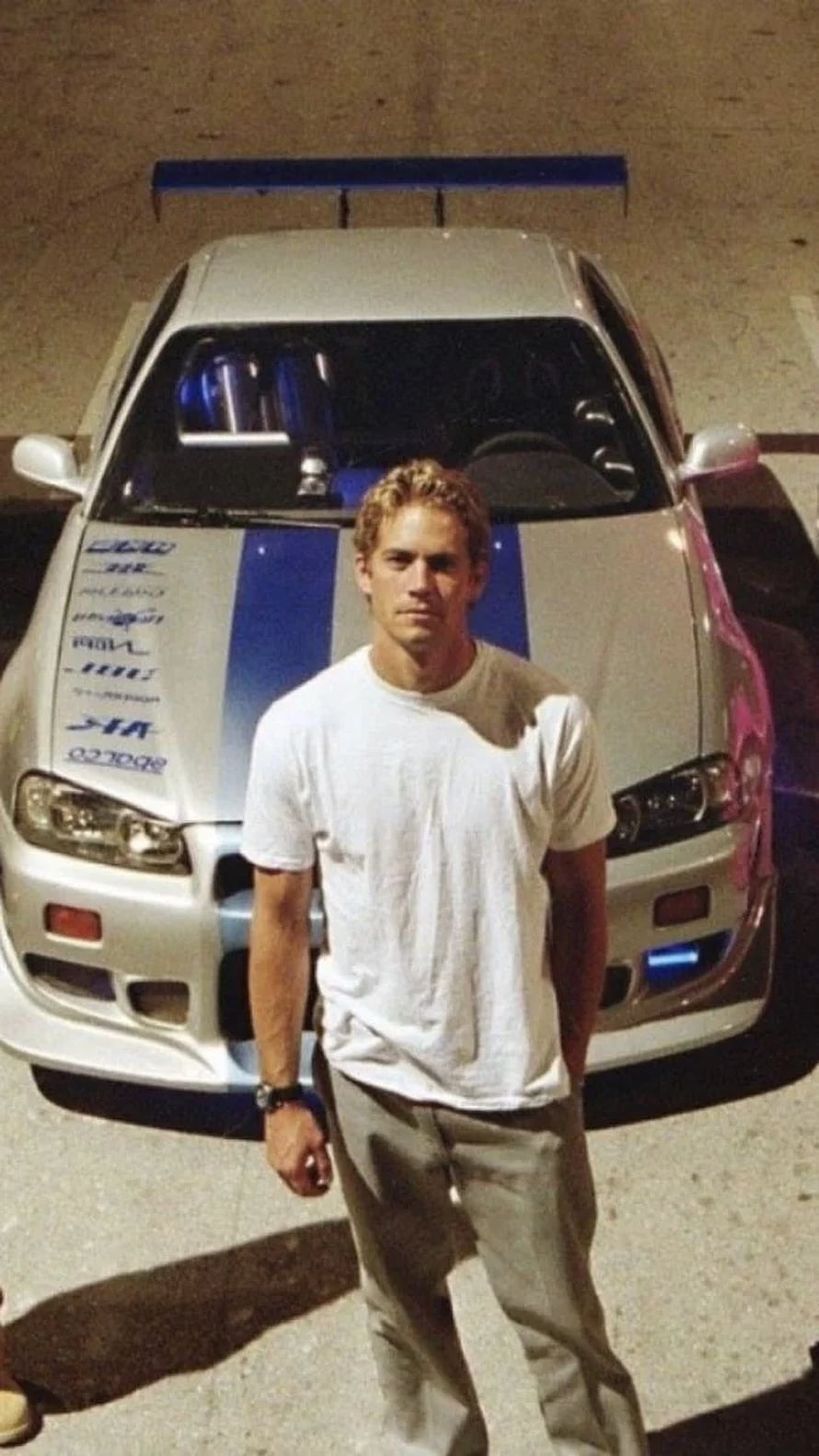 Paul Walker standing in front of a silver Nissan Skyline GT-R with blue racing stripes HD phone wallpaper