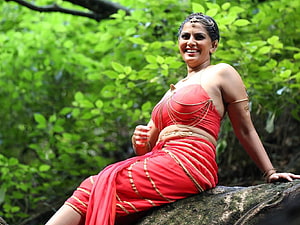 Varalaxmi Sarathkumar in a red traditional costume with gold jewelry, seated on a rock in a lush green forest HD wallpaper
