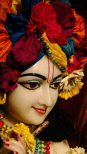Close-up of Lord Krishna statue adorned with vibrant floral headdress and traditional jewelry HD phone wallpaper