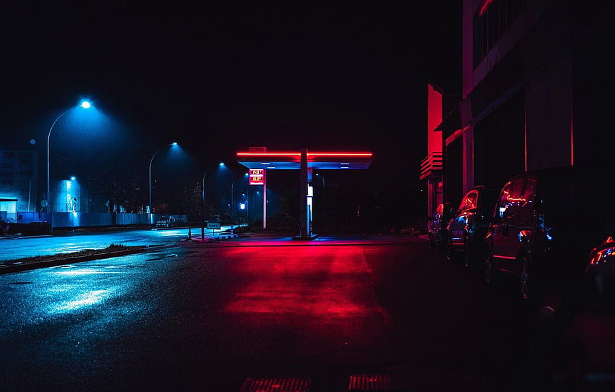 Neon-lit gas station at night with wet street reflections and parked cars HD wallpaper