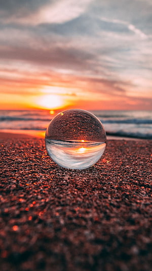 Glass lens sphere on a pebble beach capturing a vibrant ocean sunset HD phone wallpaper