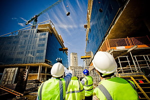 Construction workers in high-visibility vests and hard hats observing a crane lift materials at a modern building site HD wallpaper
