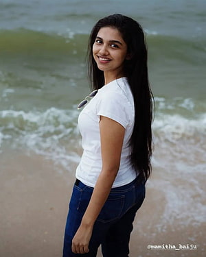 Mamitha Baiju smiling on a sandy beach wearing a white t-shirt and blue jeans HD phone wallpaper