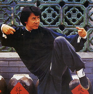 Jackie Chan in a dynamic martial arts pose wearing traditional black attire against an ornate green lattice wall HD phone wallpaper