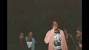 Brent Faiyaz in a pink cardigan with friends in a dark grainy night scene HD wallpaper