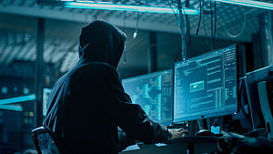 Silhouette of a hooded figure working at a multi-monitor cyber workstation in a dark room HD wallpaper