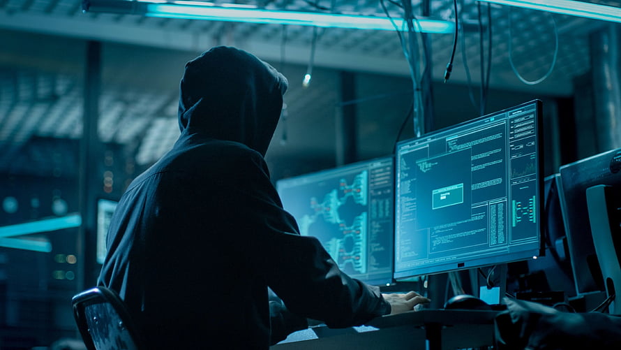 Silhouette of a hooded figure working at a multi-monitor cyber workstation in a dark room HD wallpaper