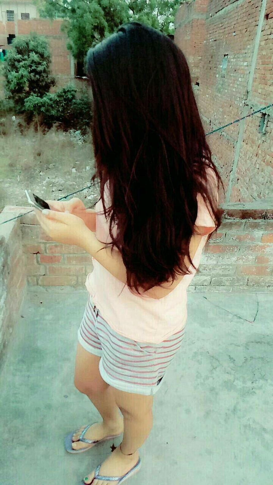 Young woman with long dark hair in pink top and striped shorts using a mobile phone outdoors HD phone wallpaper