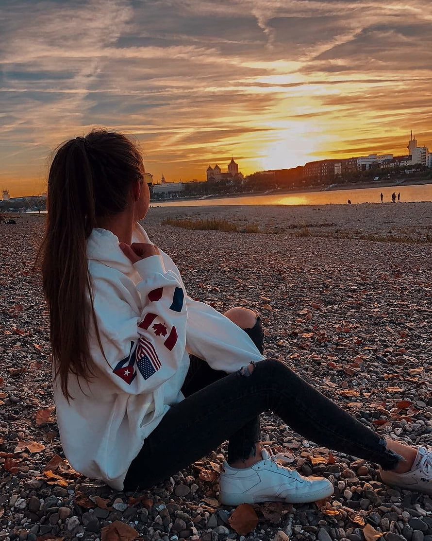 Woman in white hoodie sitting on pebble beach watching golden sunset over city skyline, hidden face poses for girls, girls hidden face HD phone wallpaper