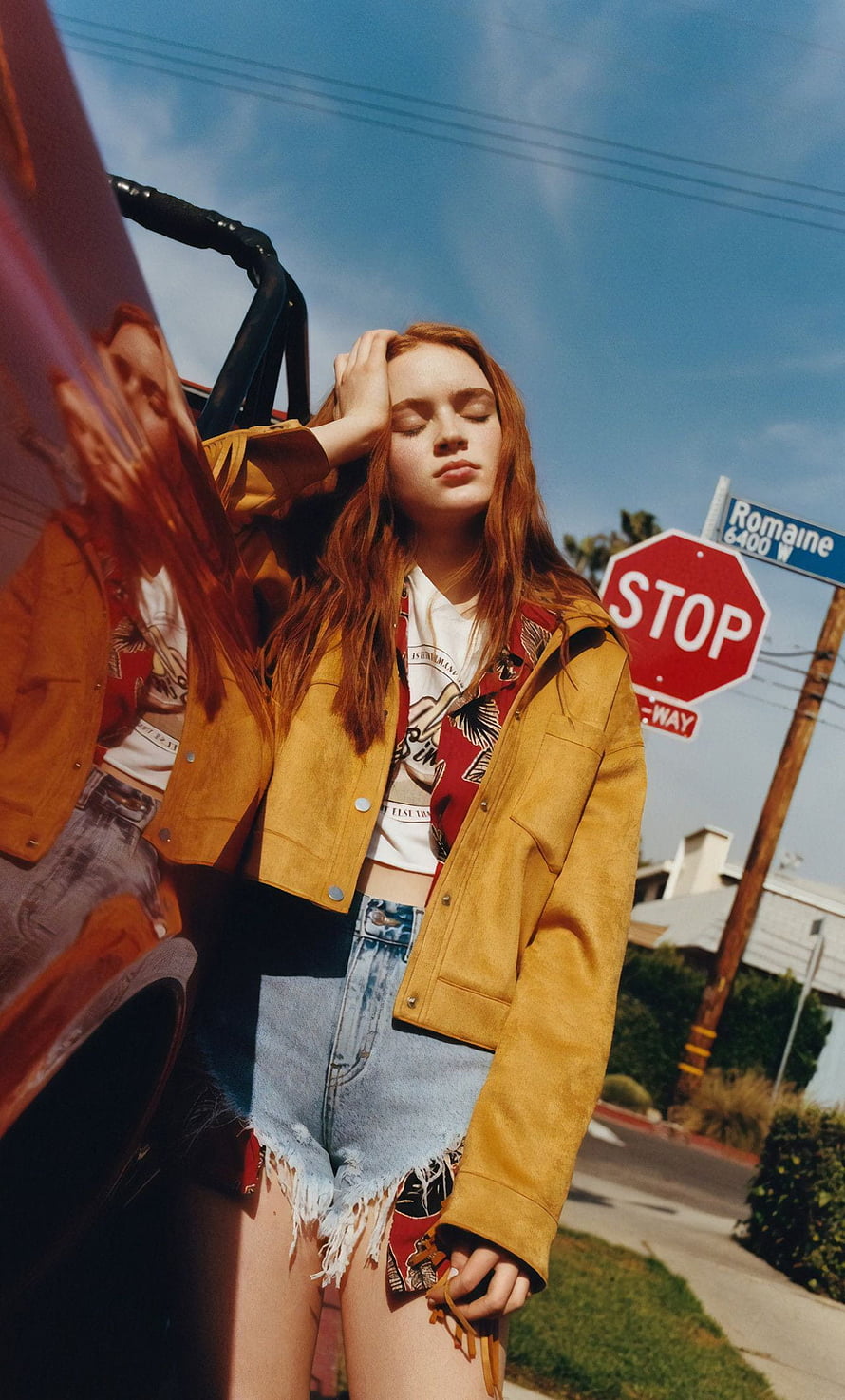 Sadie Sink in mustard jacket and denim shorts leaning against a car on a sunny street HD phone wallpaper