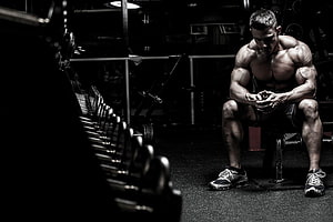 Muscular Athlete Resting on Bench in Dark Gym with Dumbbells HD wallpaper